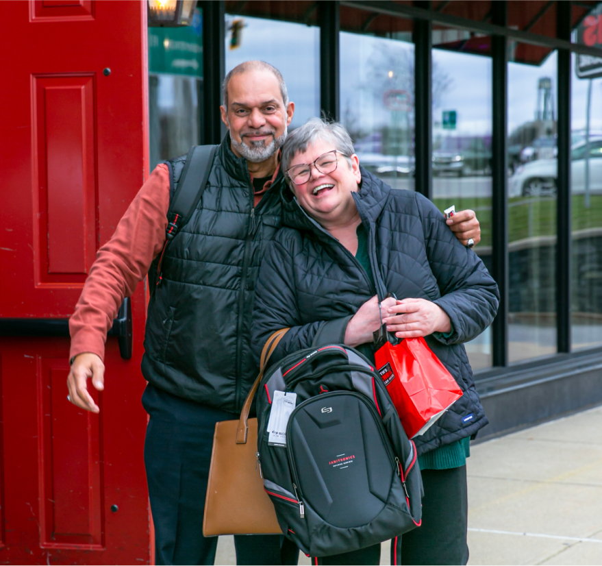 A happy man and woman pose together, holding bags and backpacks in a casual setting