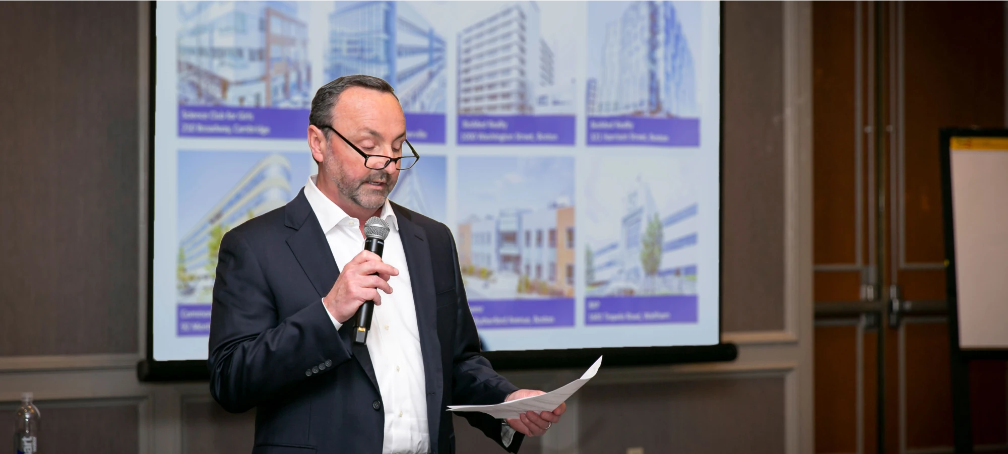 Professional speaker in glasses delivers a presentation with architectural renderings displayed behind him