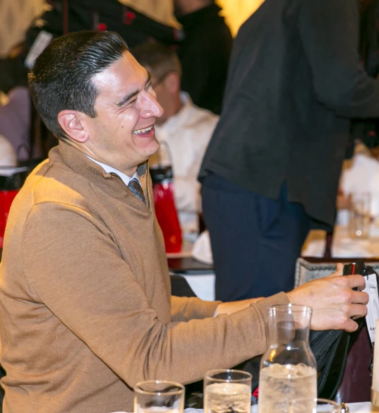 Man in a tan sweater laughs while seated at a crowded event with gift bags on tables