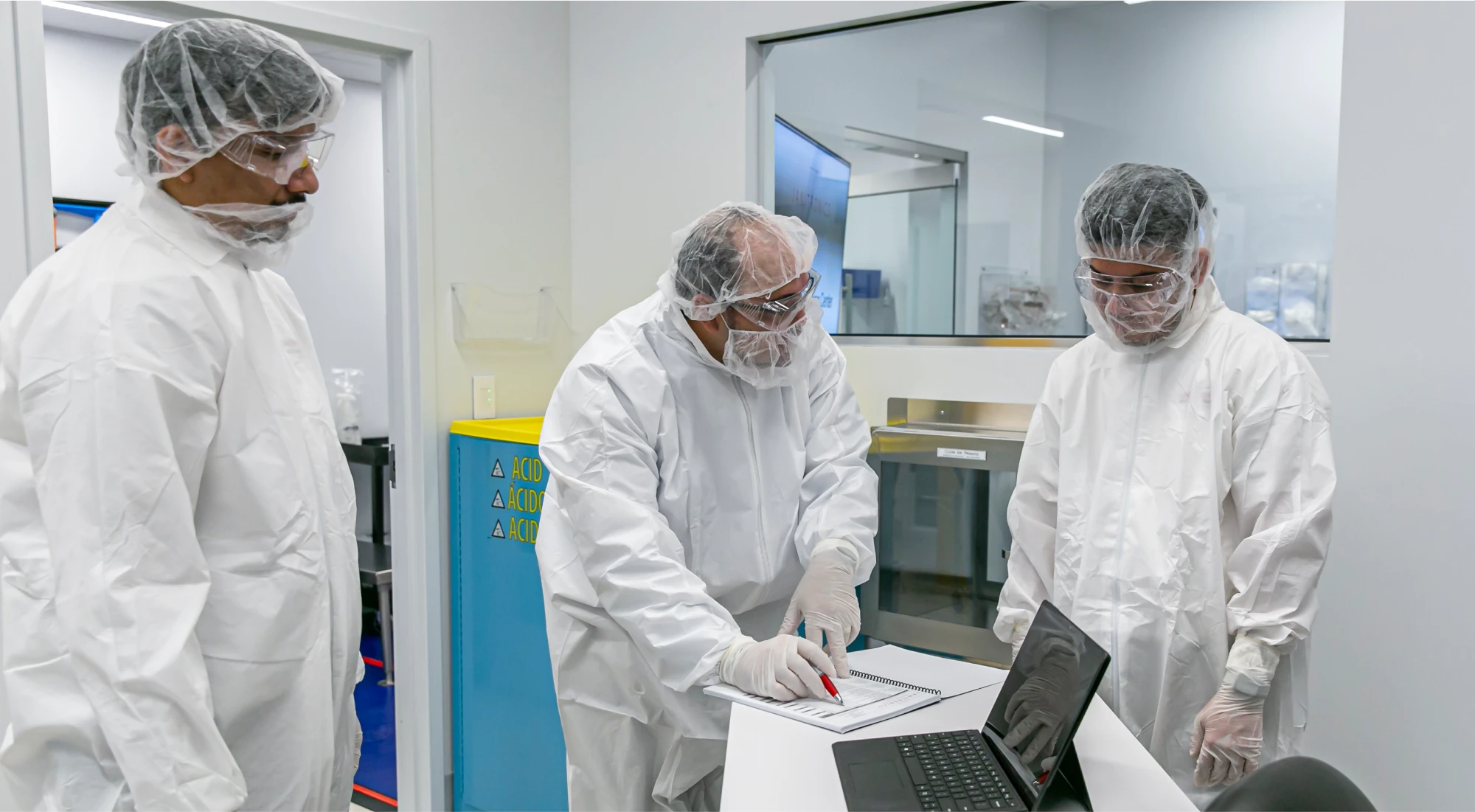 Three lab technicians wearing full protective suits reviewing documents in a sterile cleanroom environment