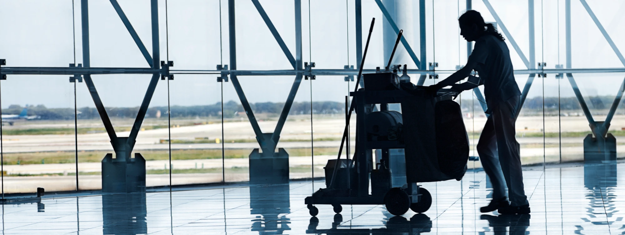 Silhouette of a janitorial worker pushing a cleaning cart in a large airport terminal