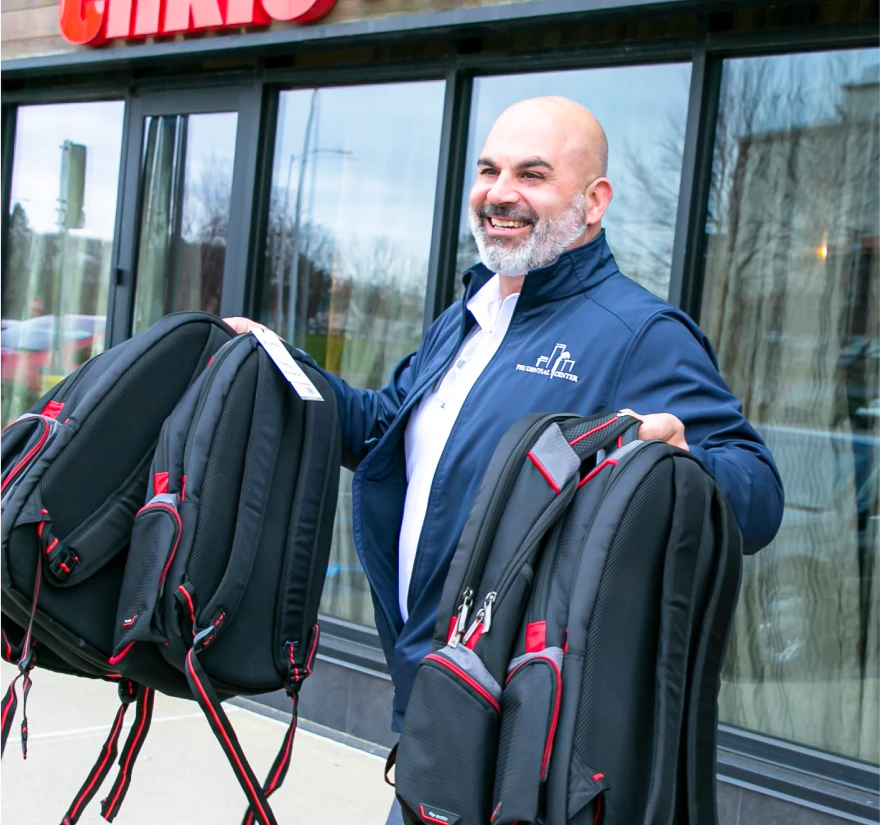 Smiling man holds multiple black and red backpacks outside a restaurant entrance