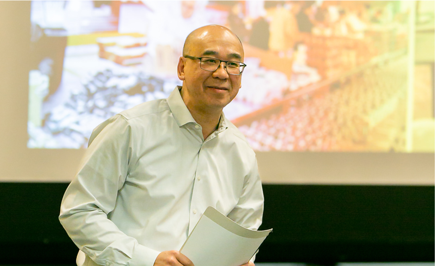 Smiling man in glasses holds papers while presenting in front of a large projection screen