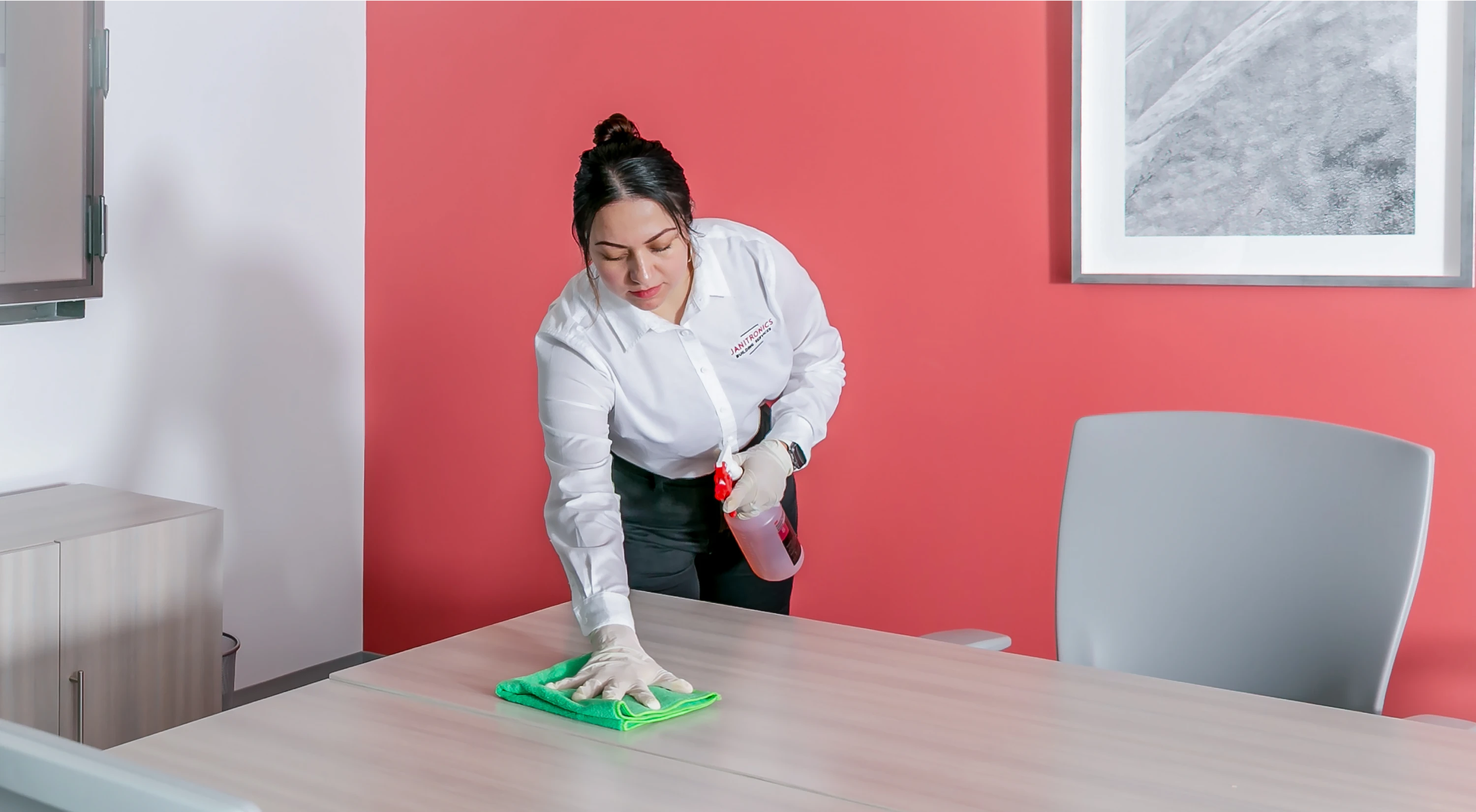 Female janitor in uniform cleaning a conference table with a green microfiber cloth and spray bottle