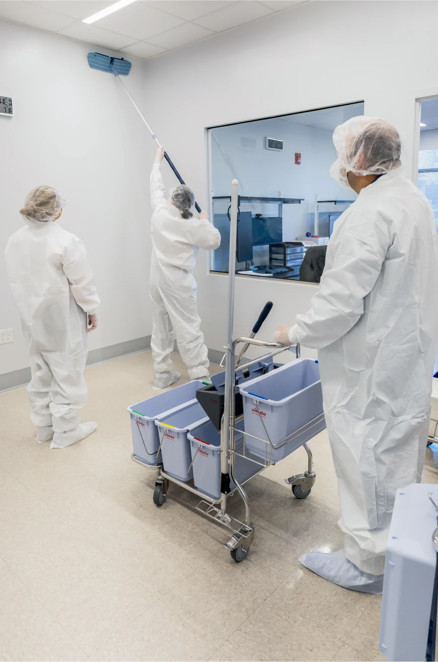 Workers in sterile white coveralls sanitizing a laboratory wall using specialized cleaning tools
